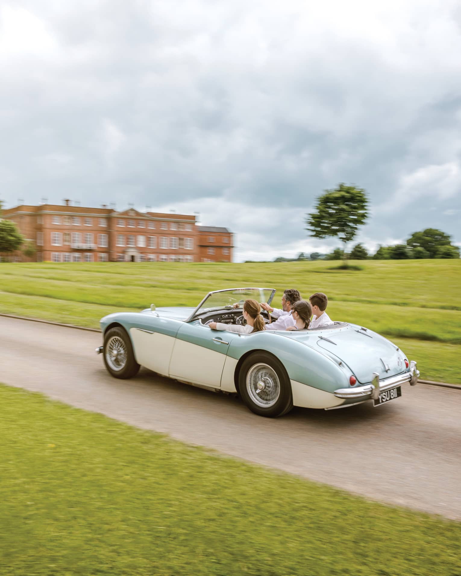 Family rides in vintage convertible car down road past English countryside to hotel manor