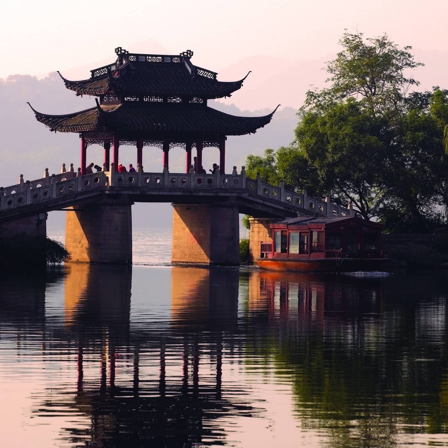 A boat floats towards a stone bridge with a pagoda cover cast in orange light from the setting sun.