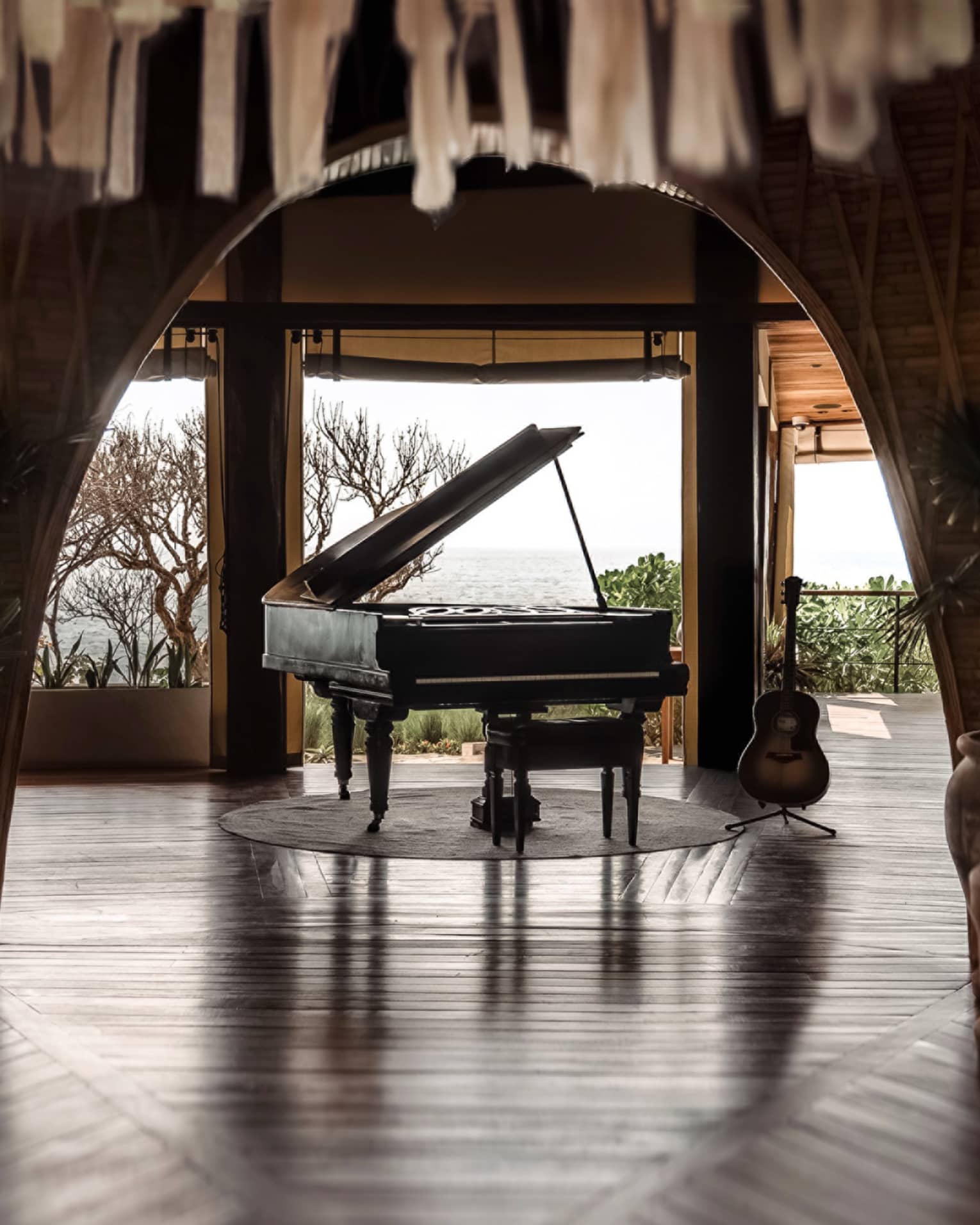 Black grand piano sits in the centre of a room that opens up to the ocean in the distance