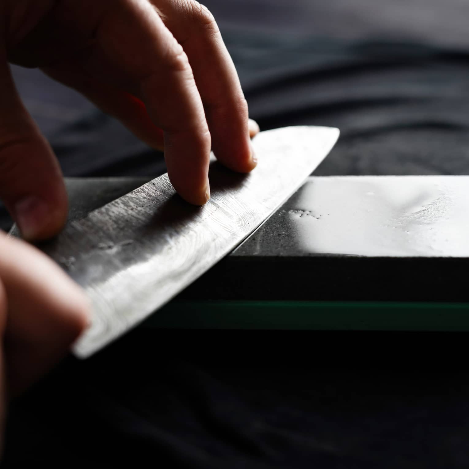 Close-up of two hands holding a chef’s knife against a gleaming dark-grey sharpening stone on a dark surface.