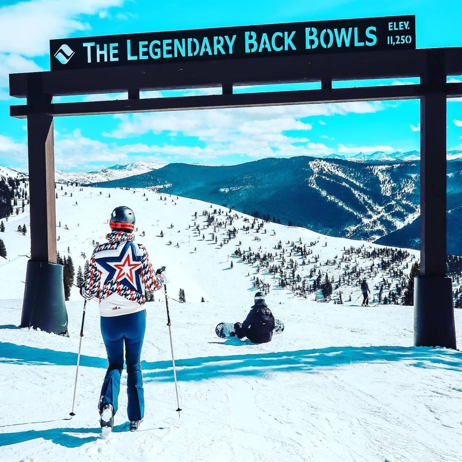 ,A woman on skiis stands on a slope beneath a wooden sign that reads "The Legendary Back Bowls"