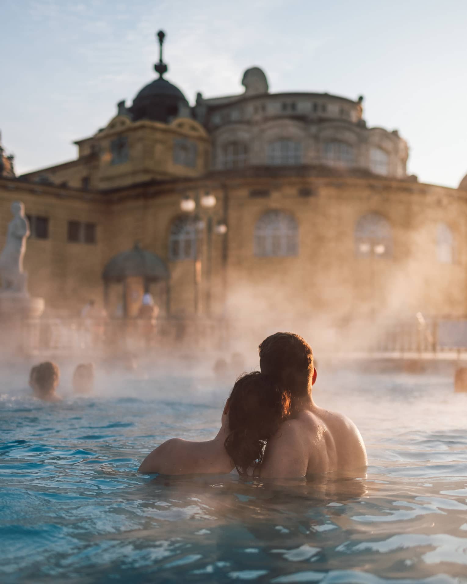 Couple huddled together in thermal pool at Szechenyi Thermal Baths in Budapest, other bathers in background