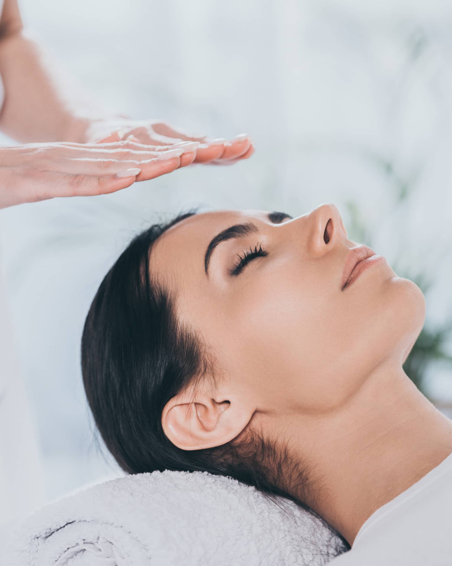 Woman with eyes closed lays on rolled up towel on spa table under therapists' hovering hands