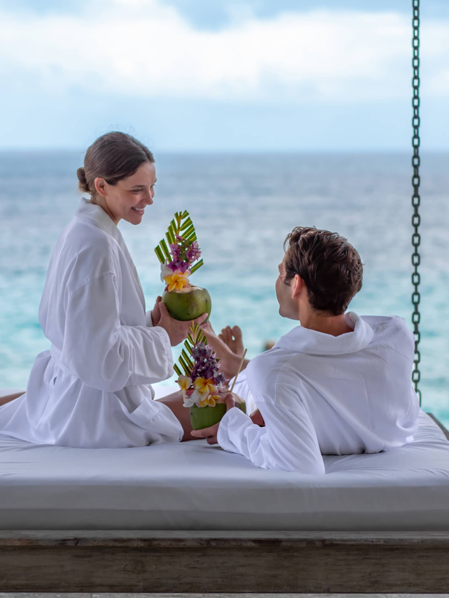 couple enjoying a sea view and a cocktail on a hanging beach bed whilst wearing white robes
