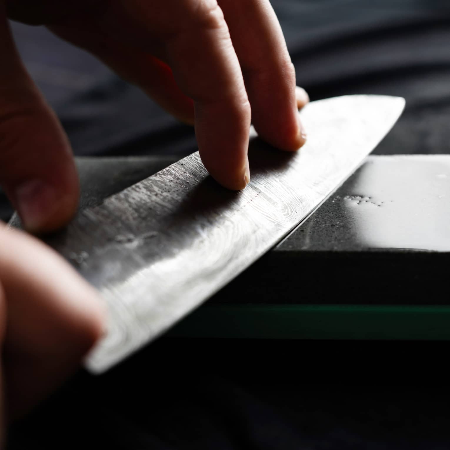 Close-up of two hands holding a chef’s knife against a gleaming dark-grey sharpening stone on a dark surface.