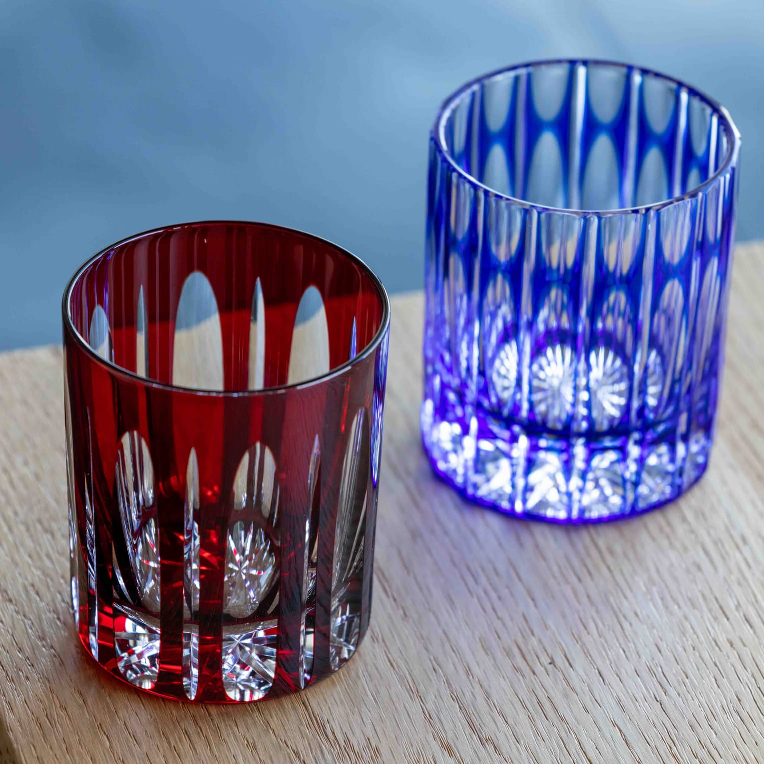 Close-up of two cut-glass tumblers with intricate geometric patterns overlaid by coloured glass, one dark red, one blue.
