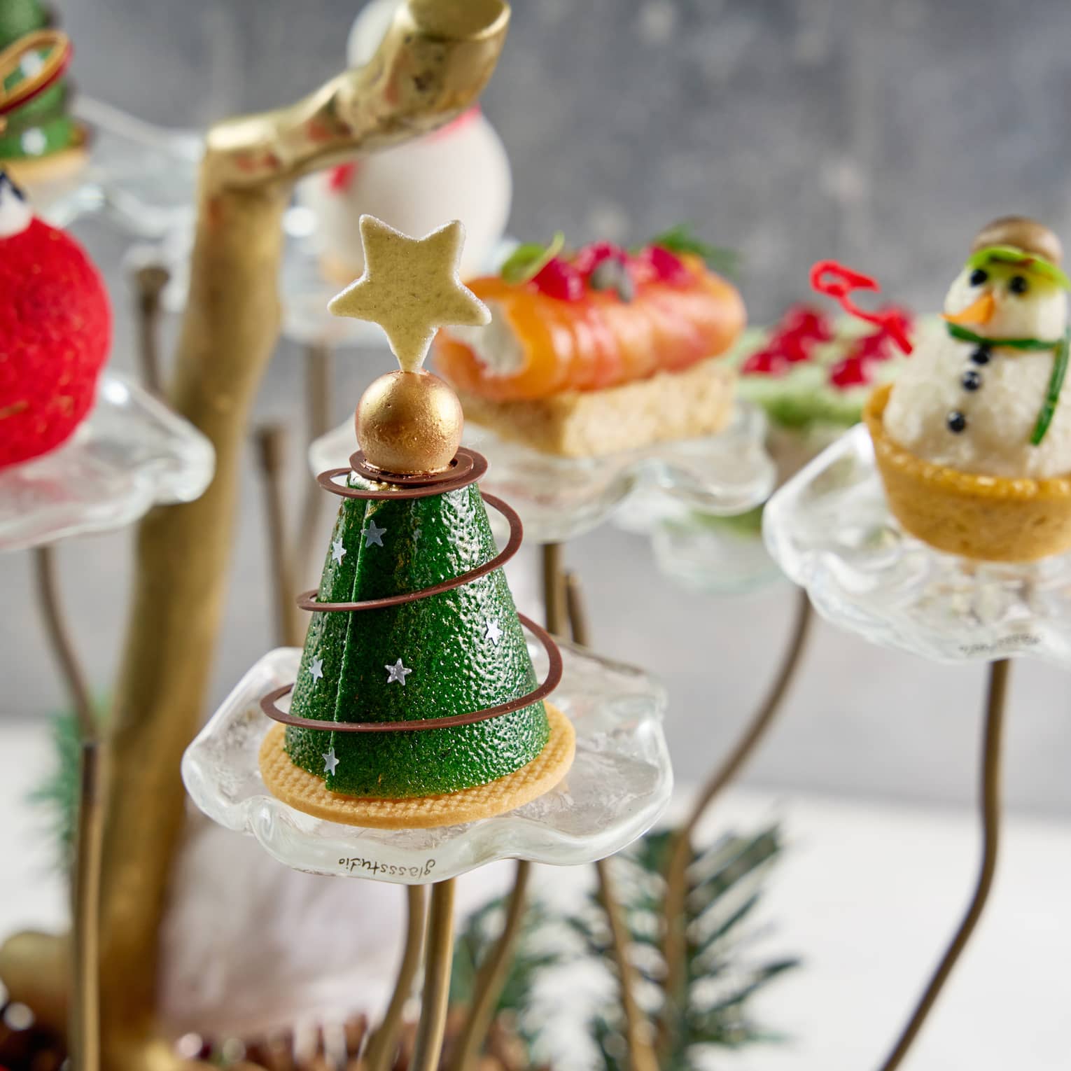 Holiday pastries shapped like a  Christmas tree, snowman, red ball ornament and small house are on display on small glass pedastals