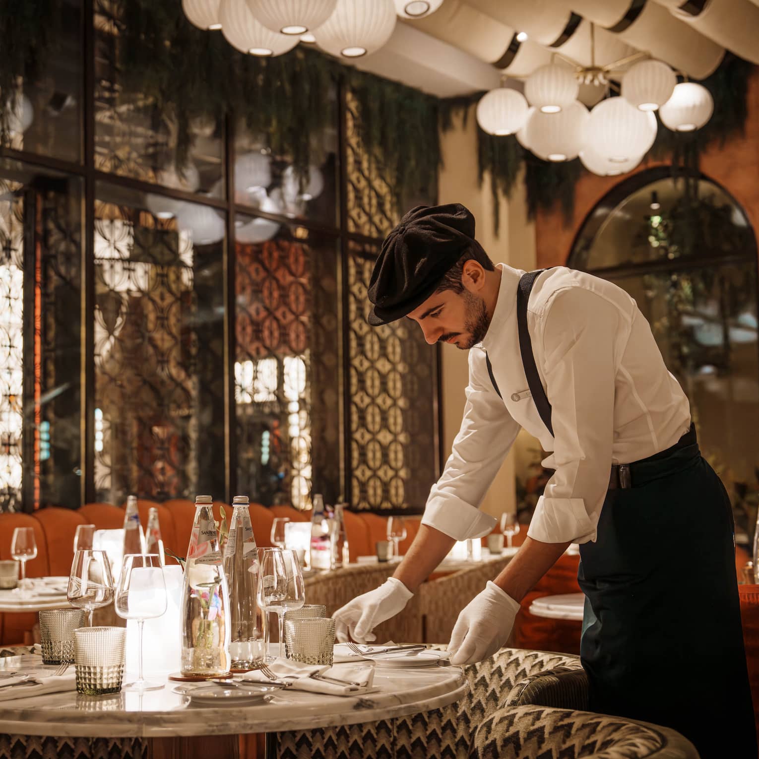 Waiter wearing white button-down shirt, black pants, black suspenders, black beret and white gloves arranges a plate on a table in an Art Deco?style restaurant