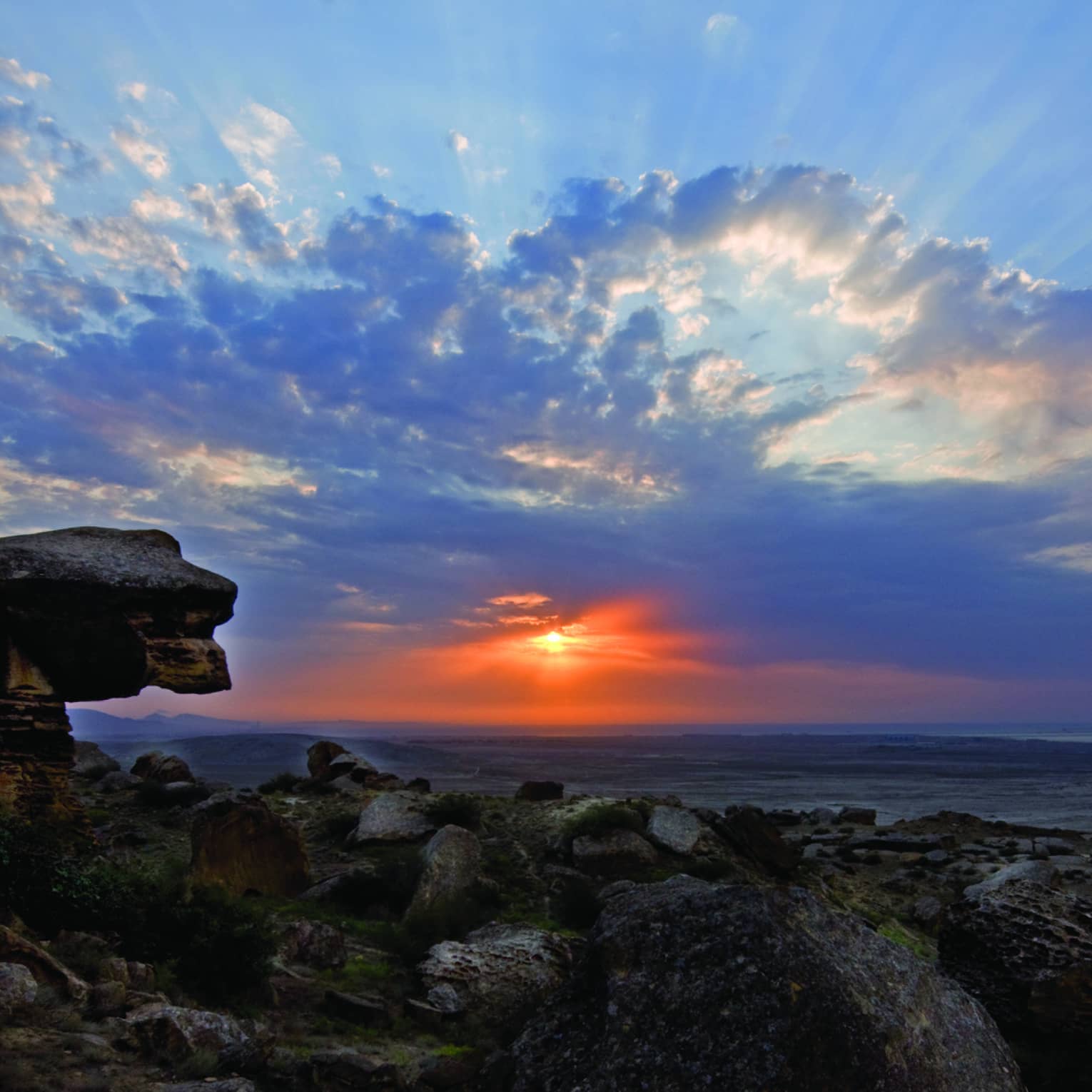 Sunbeams and fluffy azure blue clouds dance across a twilight sky, the vibrant red sun gently illuminating a rocky shoreline.