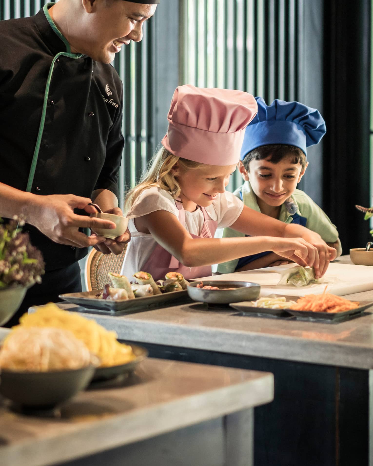 Chef watches as children wearing tall chef hats make spring rolls at counter