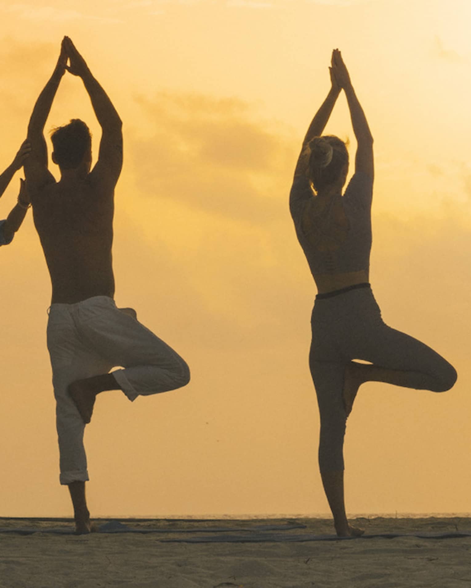 The morning sun silhouetting a couple doing yoga poses with an instructor on a beach surrounded by palm trees and greenery.