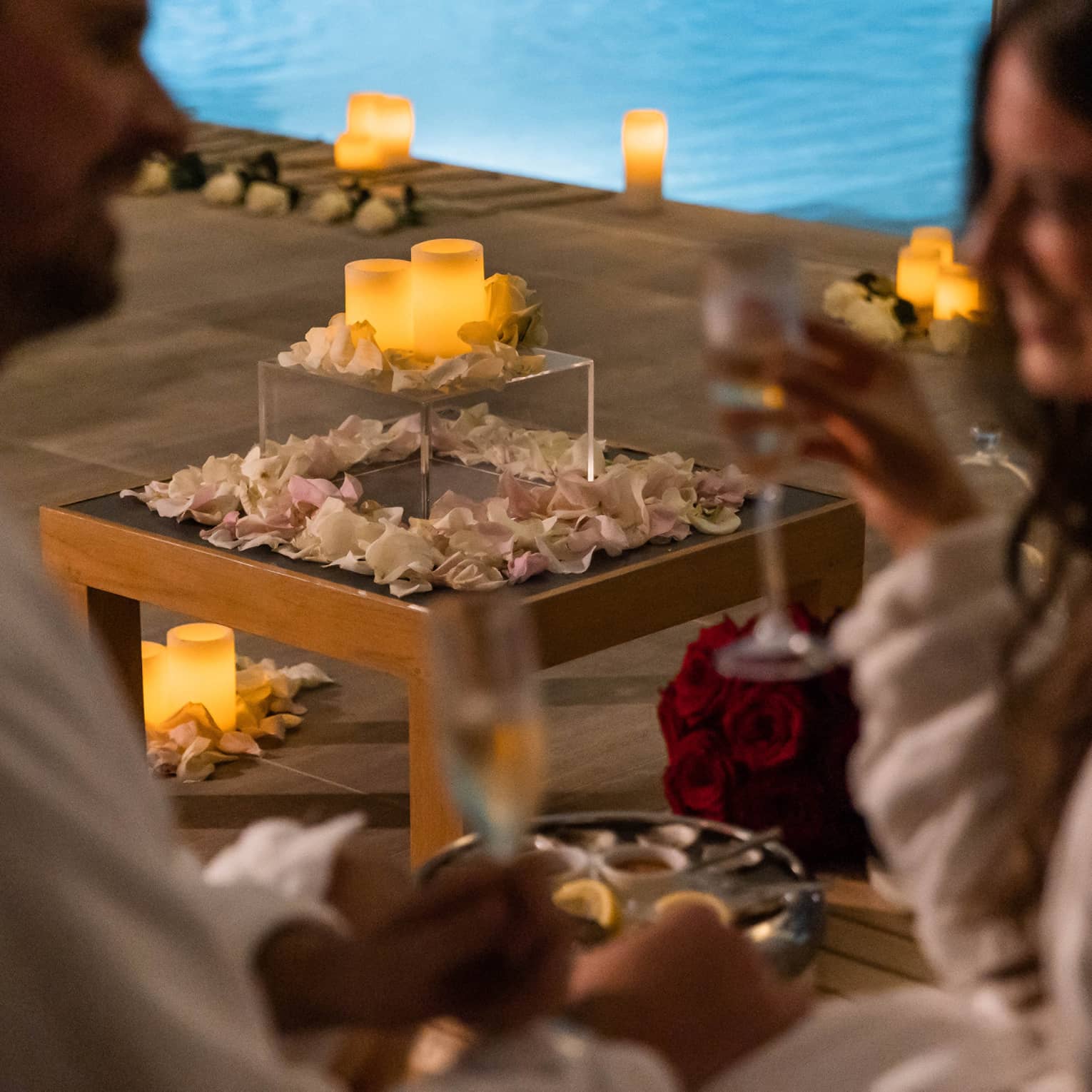 A couple with wine in hand near a pool surrounded by flowers petals and candles.