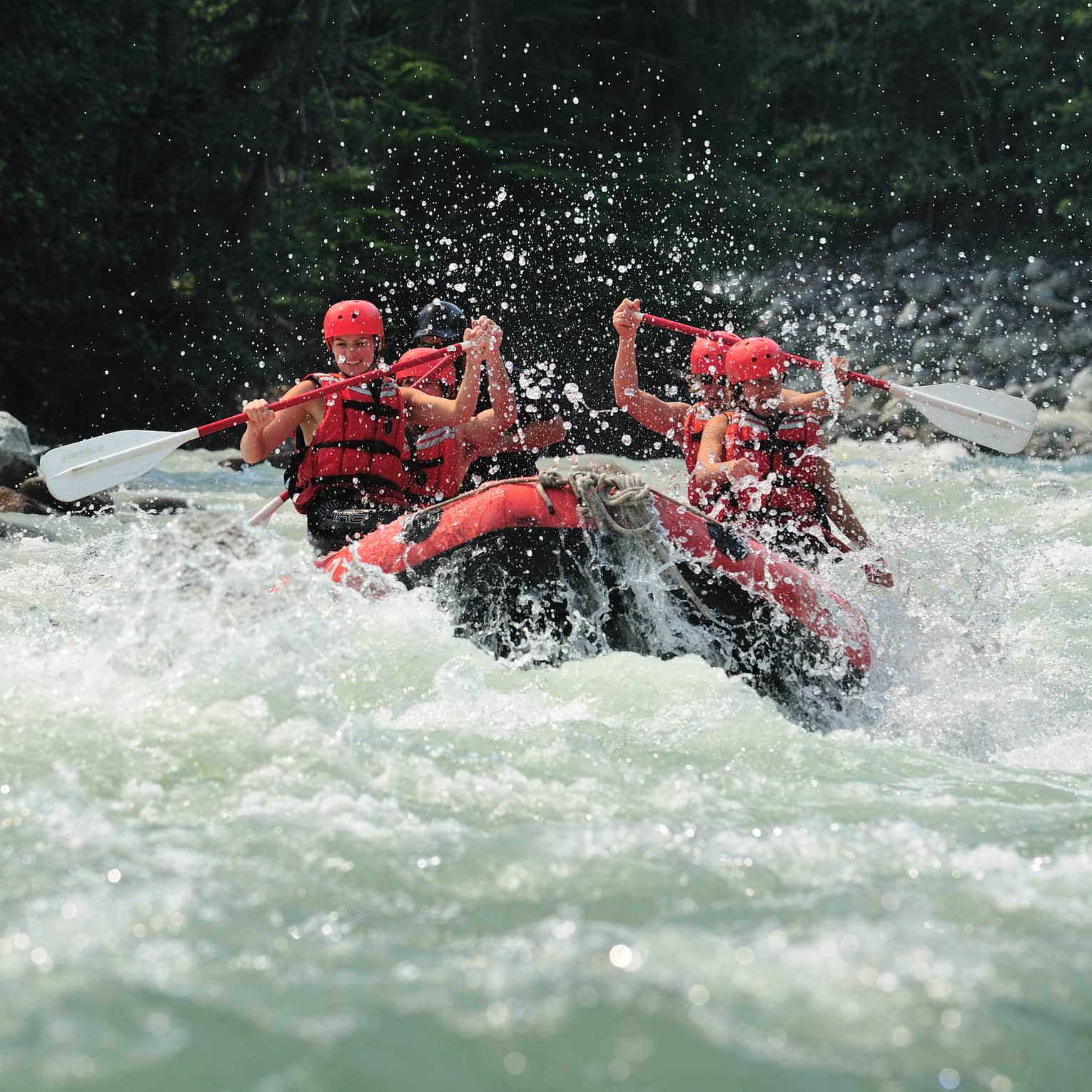 Smiling paddlers in red helmets and lifejackets steer an inflated raft through raging white water, passing evergreen trees.
