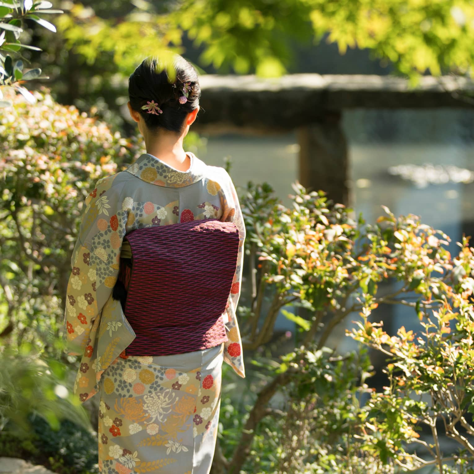 Amid bushes and trees, rear view of a person in a grey kimono with beige and orange flowers and a black and pink obi cushion.
