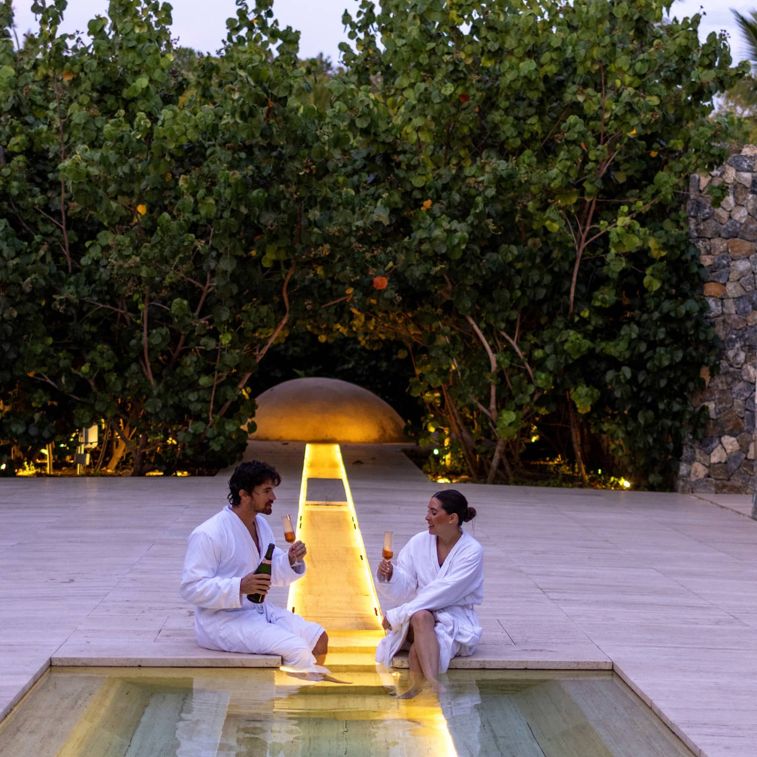 Two guests wearing spa robes and sitting by the spa tub holding wine glasses