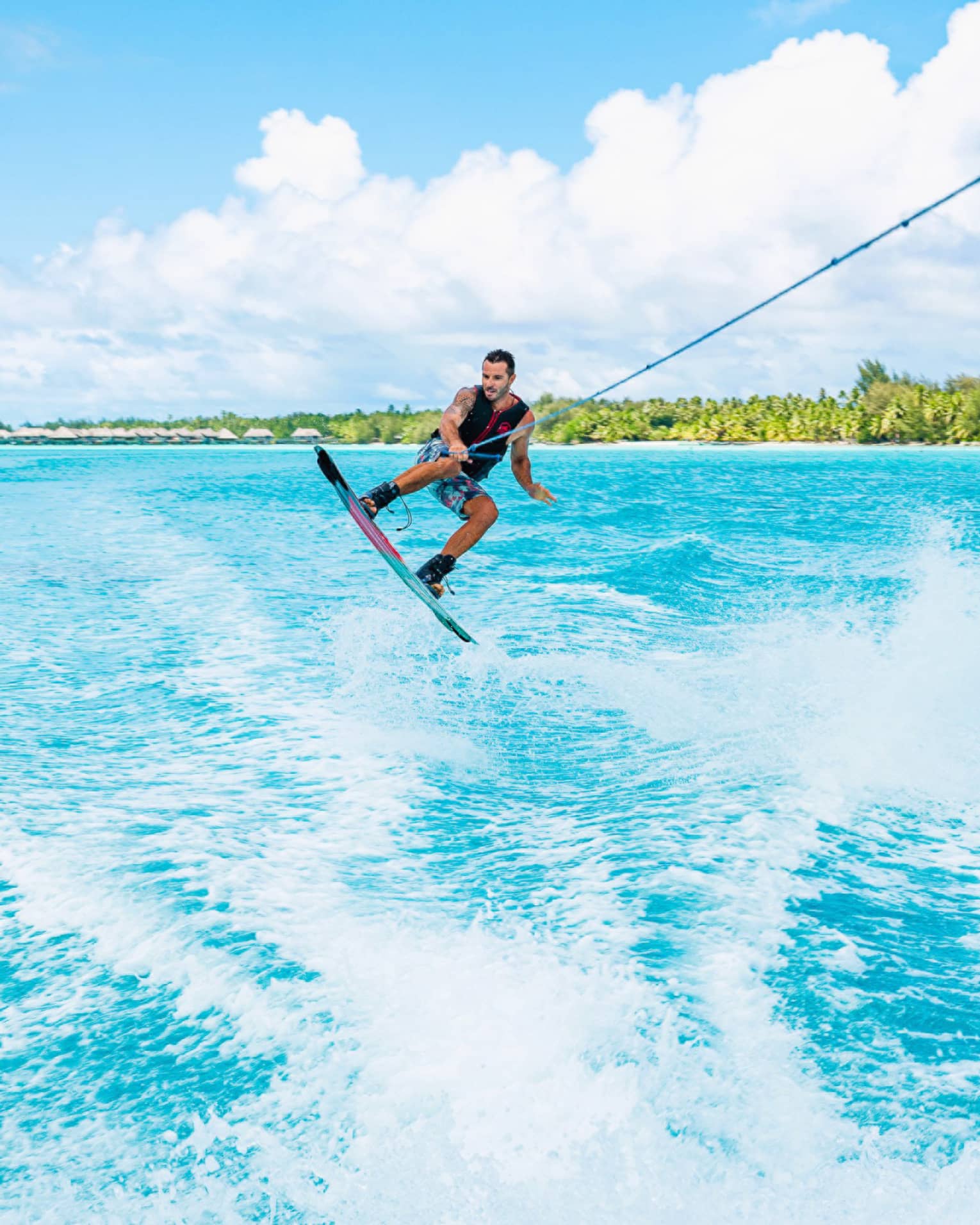 A wakeboarder leaps above the waves, one hand holding a taut rope as they're towed through the ocean beneath a sunny sky.