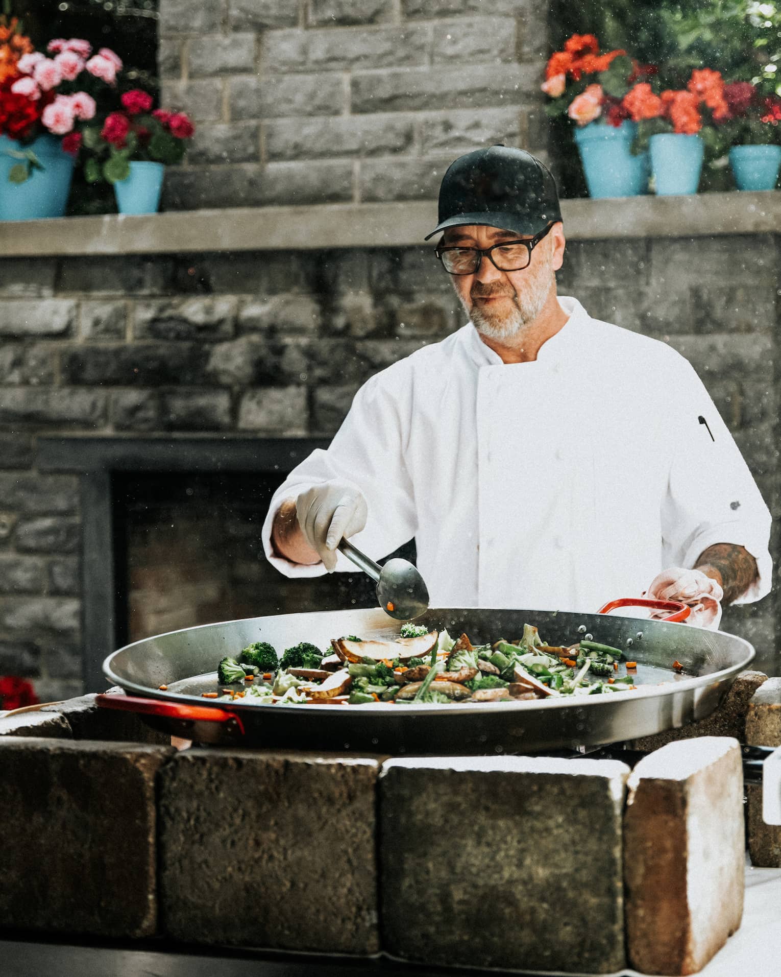 Chef wearing white chef's jacket, black ball cap and black-rimmed glasses stirs an extra-large pot of food atop an outdoor stone grill set in front of a large stone outdoor fireplace