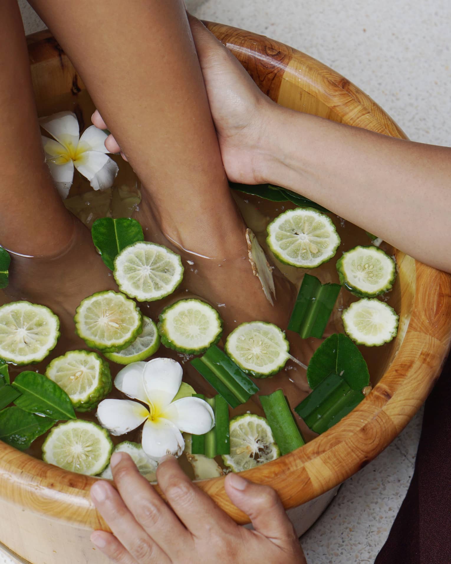 A guest receiving a foot massage at the resort spa in a wooden basin filled with water, lime slices, green leaves and white flowers.