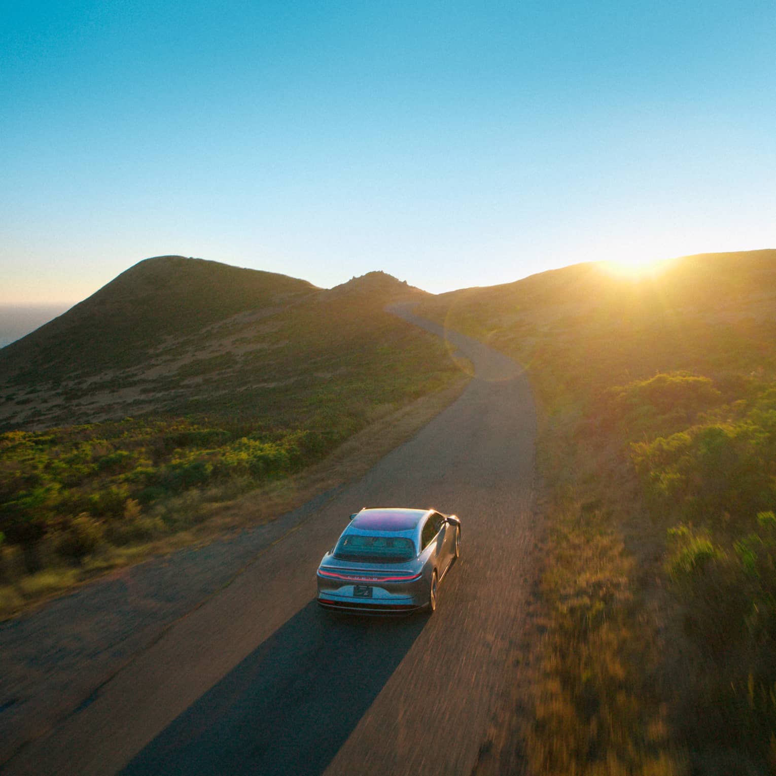 A car drives down a road surrounded by green hilly terrain. A clear blue sky with the sun just peeking over the hills is in the background, and there is also a glimpse of the ocean behind the hills.