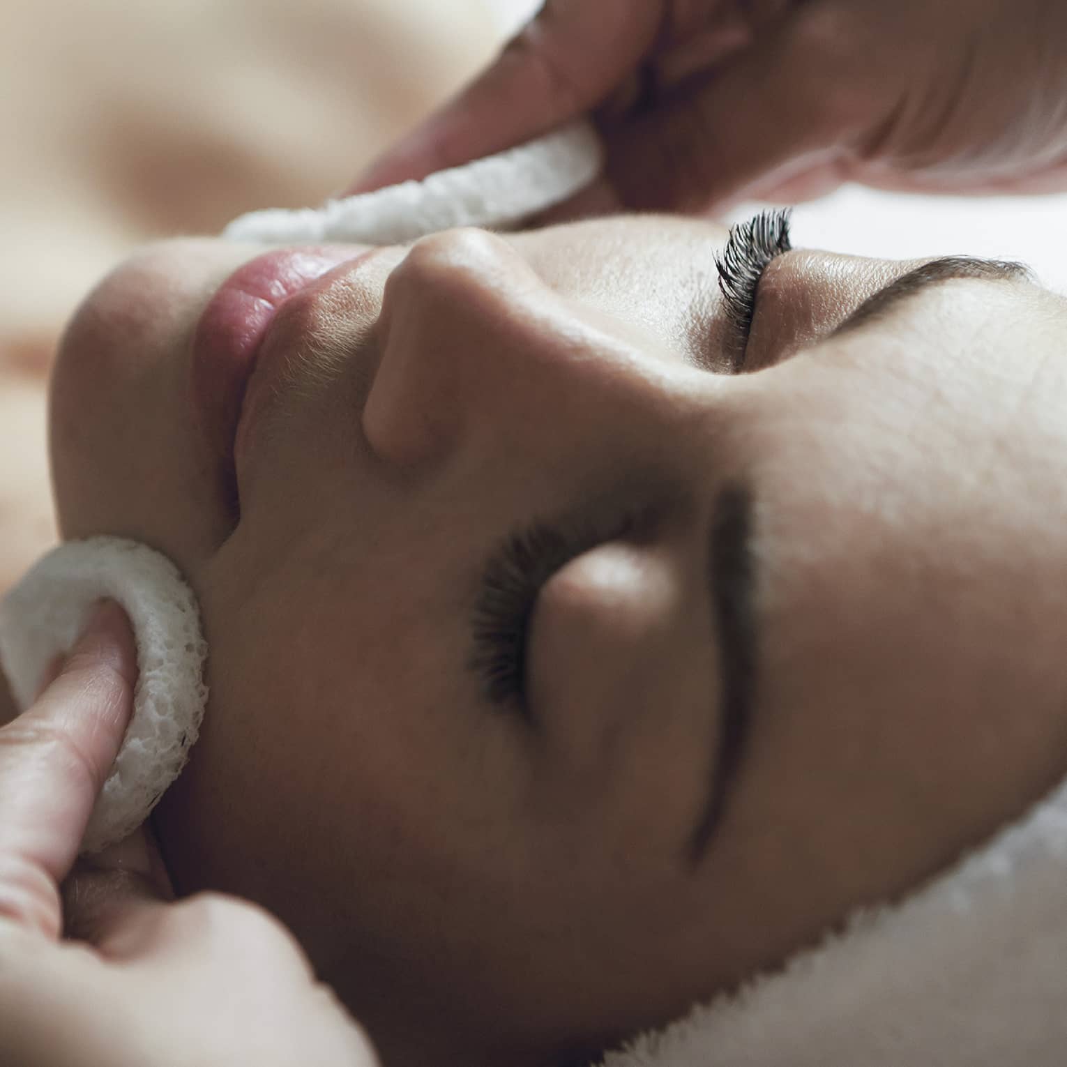 A detail of hands rubbing a sponge on a woman's face as she lies on a massage table with her eyes closed and hair wrapped up in a white towel