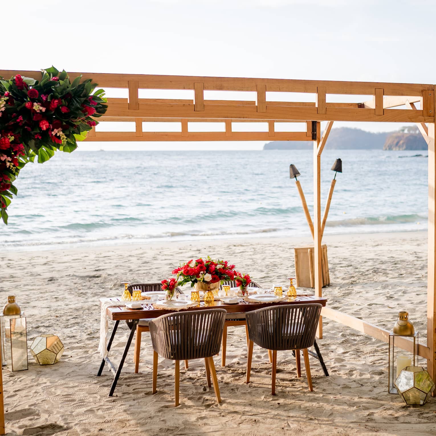 Under a pergola decorated with a vibrant bouquet of flowers, a table is set for four on the beach in the warm glow of sunset.