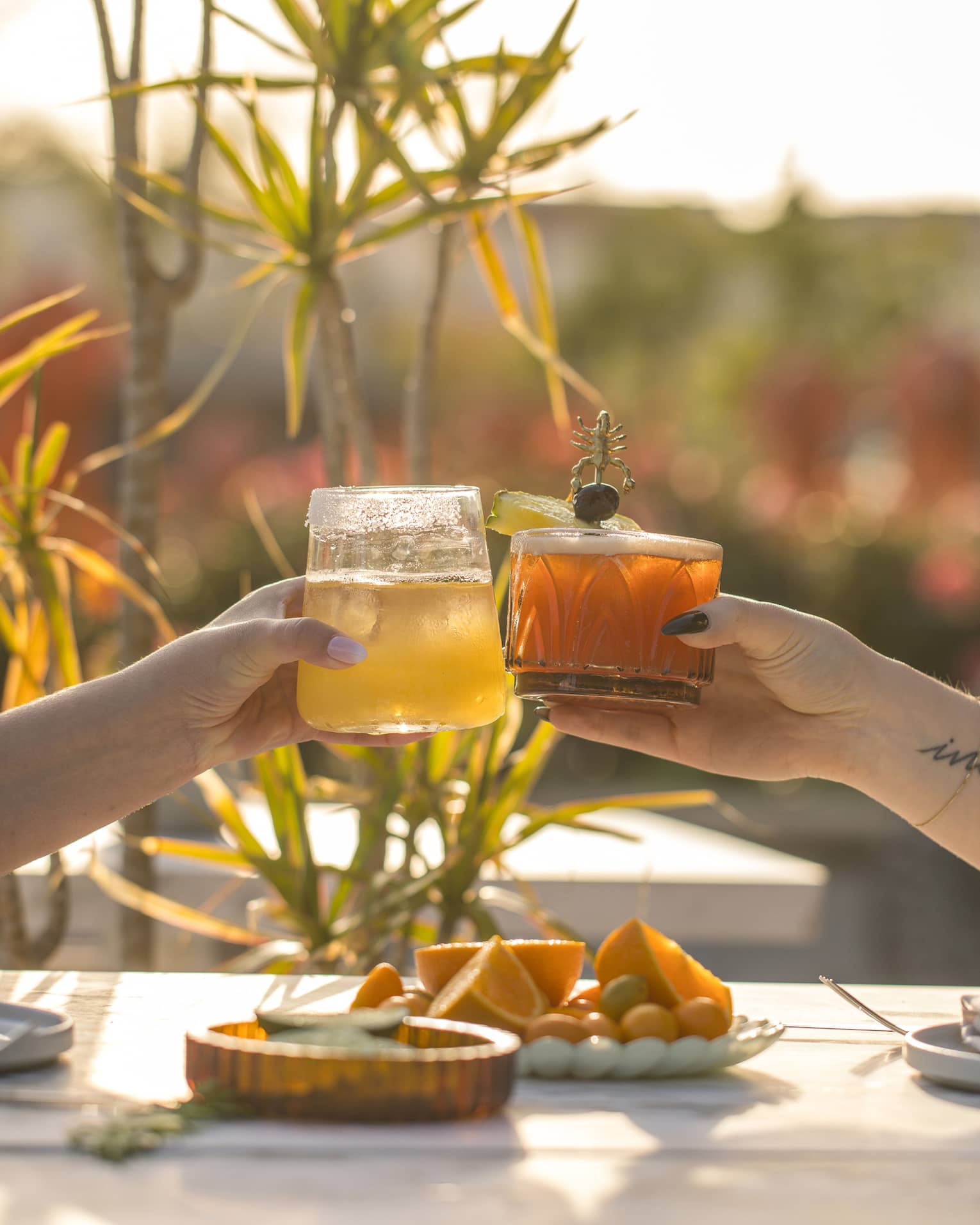 Two hands, each holding a cocktail, bring their glasses together over a sun-lit outdoor table