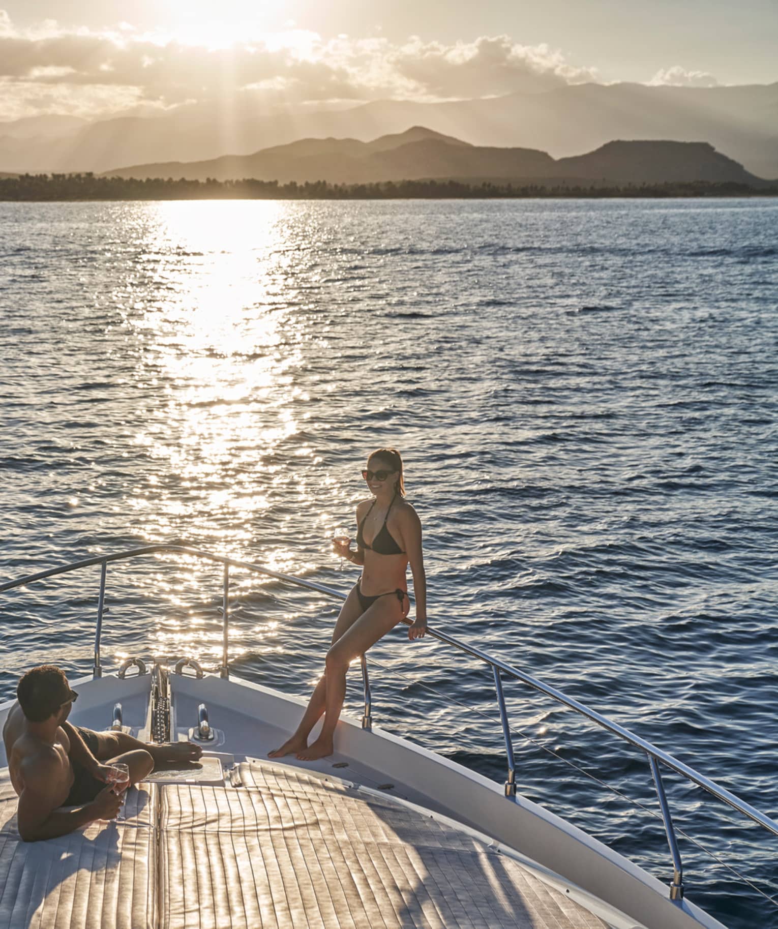 A couple in swimwear relaxing on the bow of a boat, wine glasses in hand and the setting sun glinting off the endless ocean.