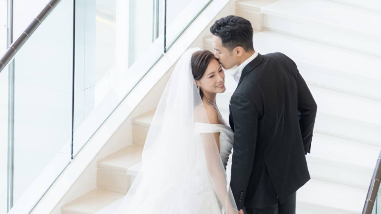 A bride and groom walking up stairs near large windows.