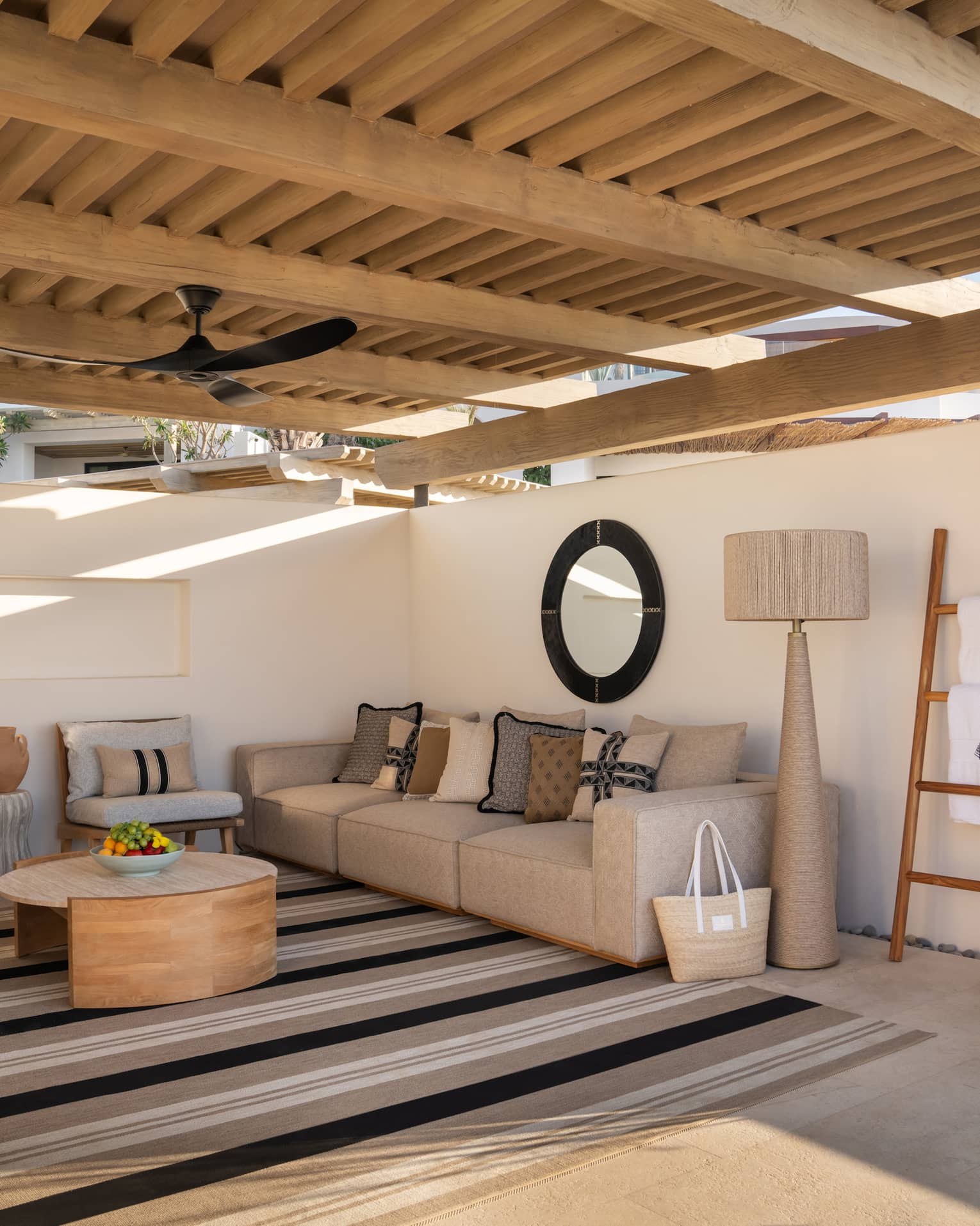 A serene cabana with a raised wooden roof and plaster walls. It's filled with comfortable furniture and a large striped rug.