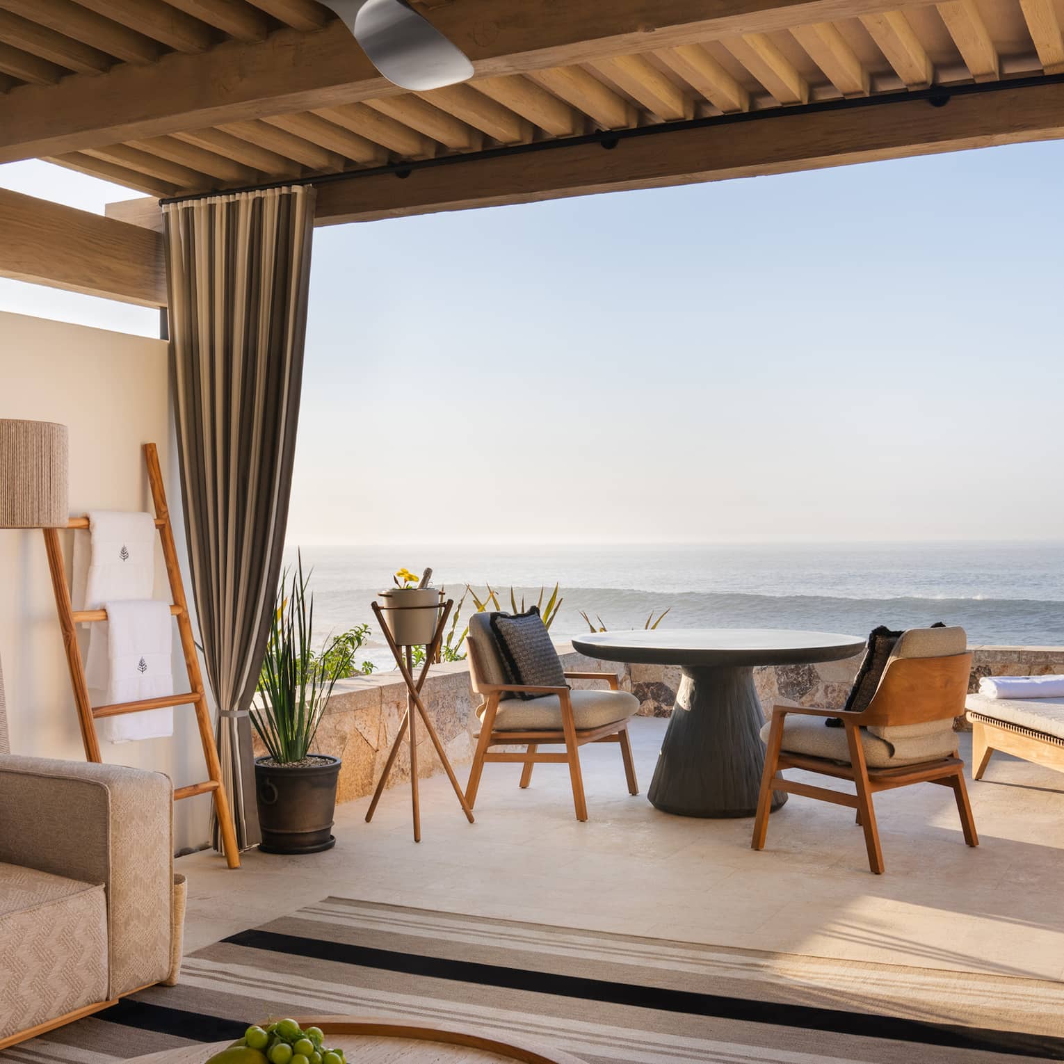 Oceanfront cabana with beige sofa, lamp and round wood coffee table under a roof with a round table and chairs and two lounge chairs out on the open-air patio overlooking the ocean