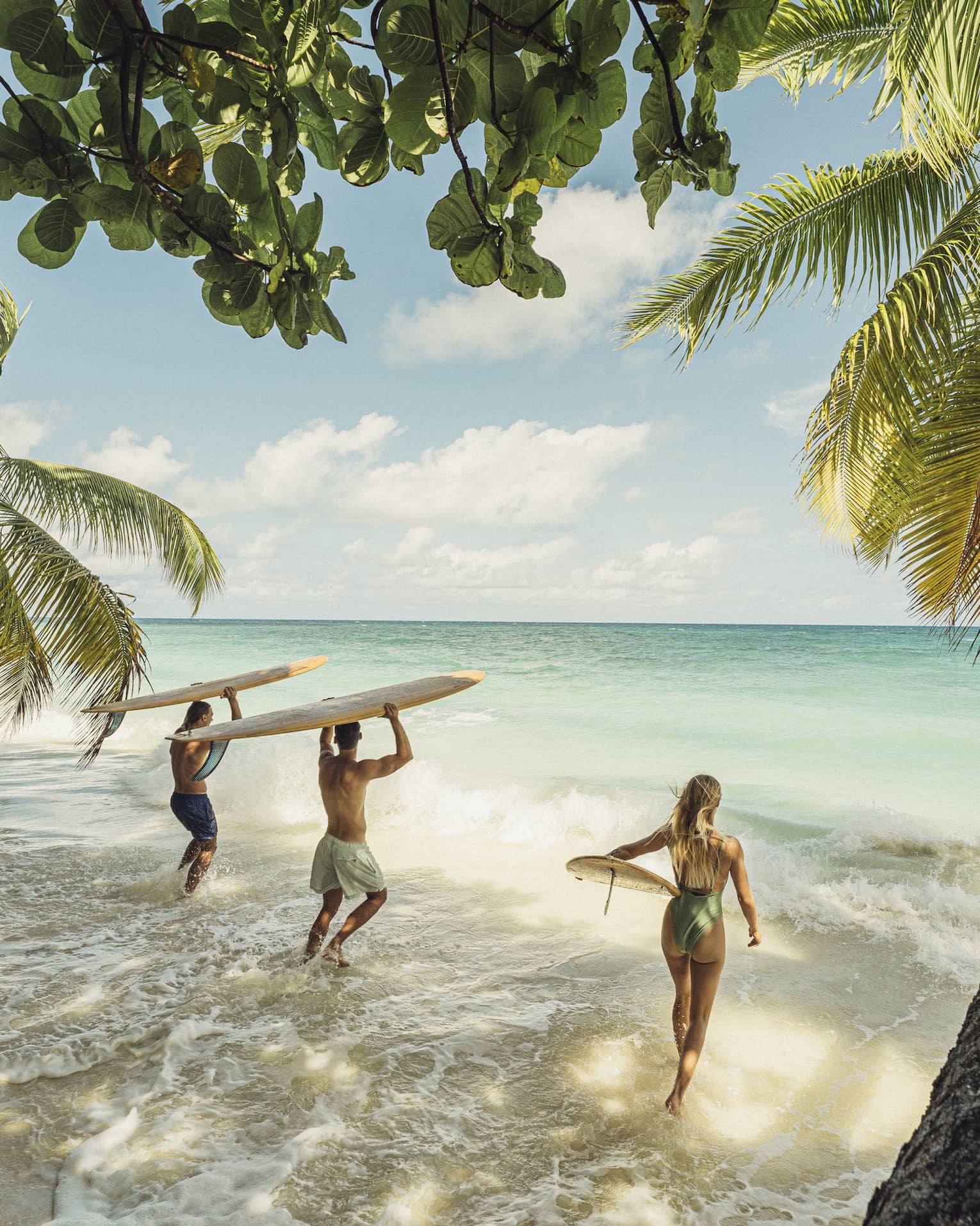 Palm trees extend over waves rushing a sandy beach, as a group of surfers with their boards bound toward the turquoise sea. 