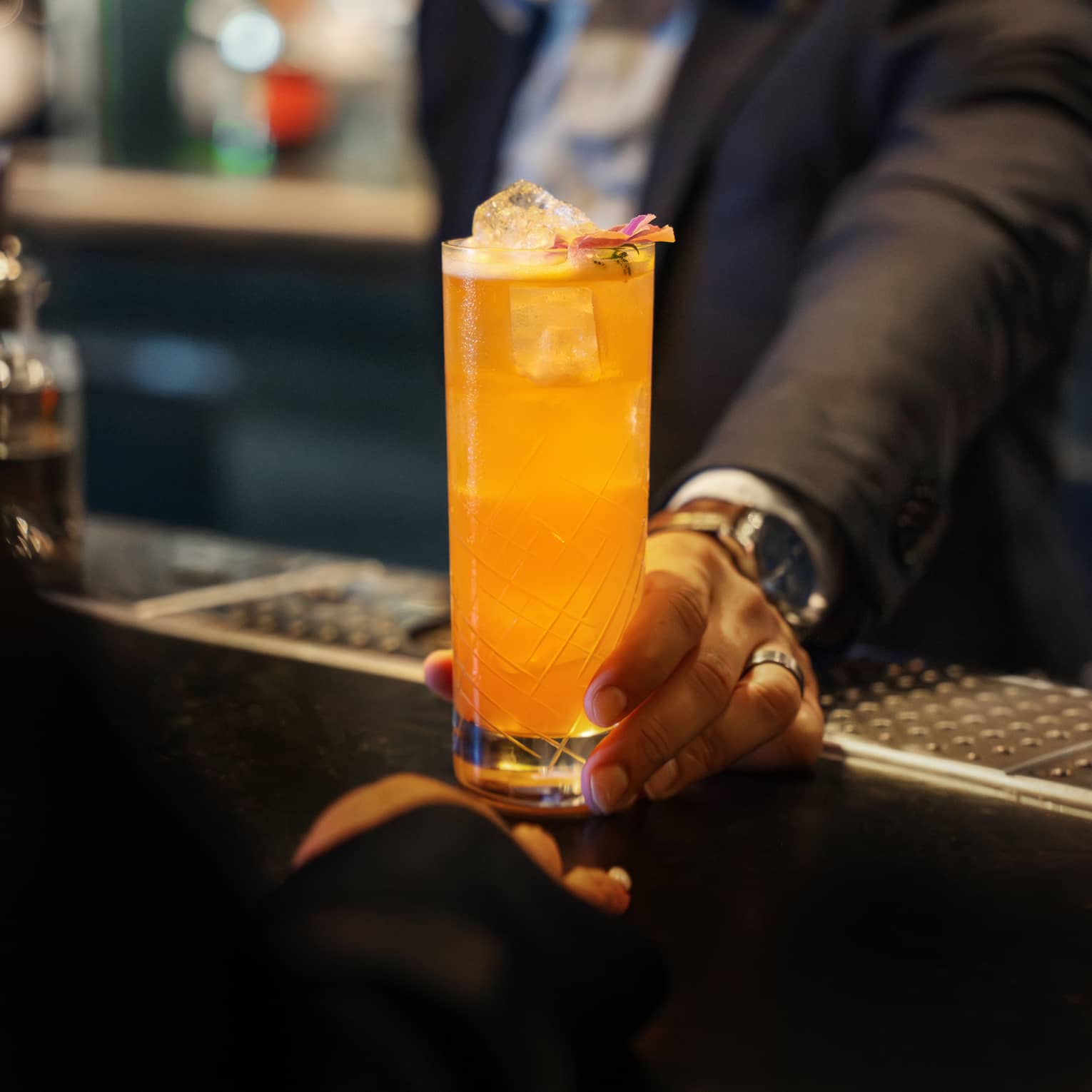 A person sets a bright orange drink in a tall clear glass on a bar counter.