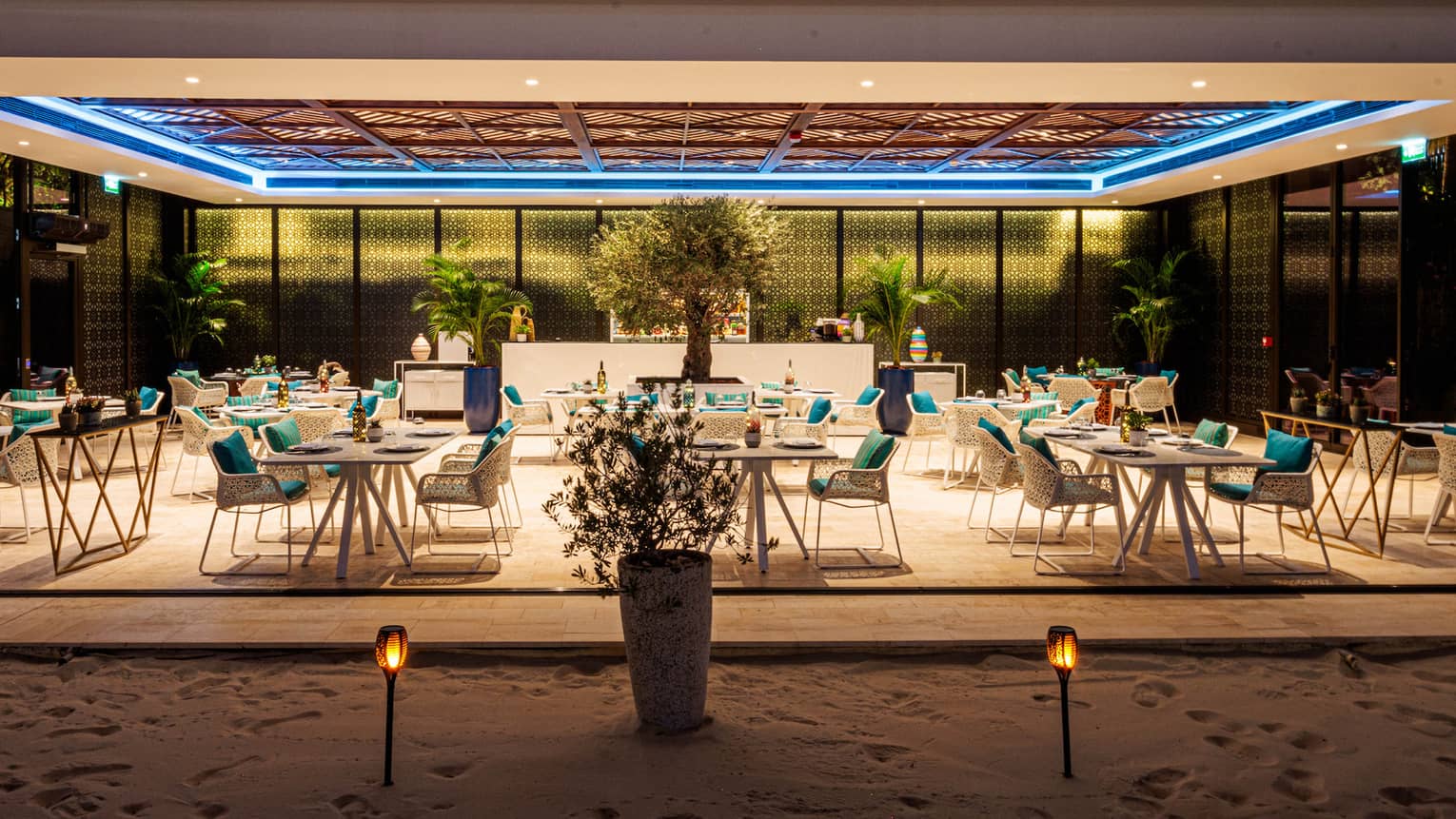 Nighttime view of restaurant dining room open to the beach on one side and surrounded by glass windows on the other sides with a tray ceiling above featuring an inset blue light