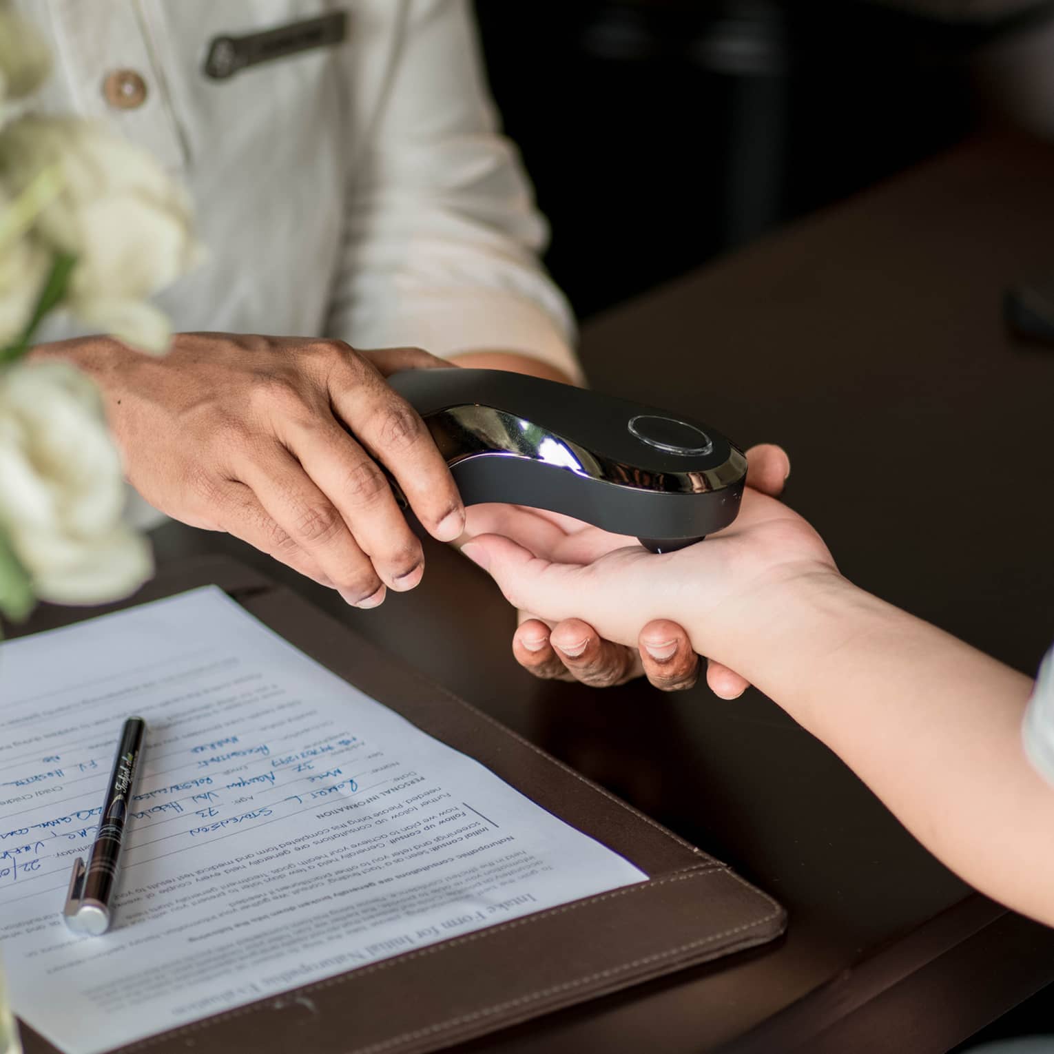 A person scans another's palm with a smooth, shiny, black electronic device beside a completed medical form on the table..