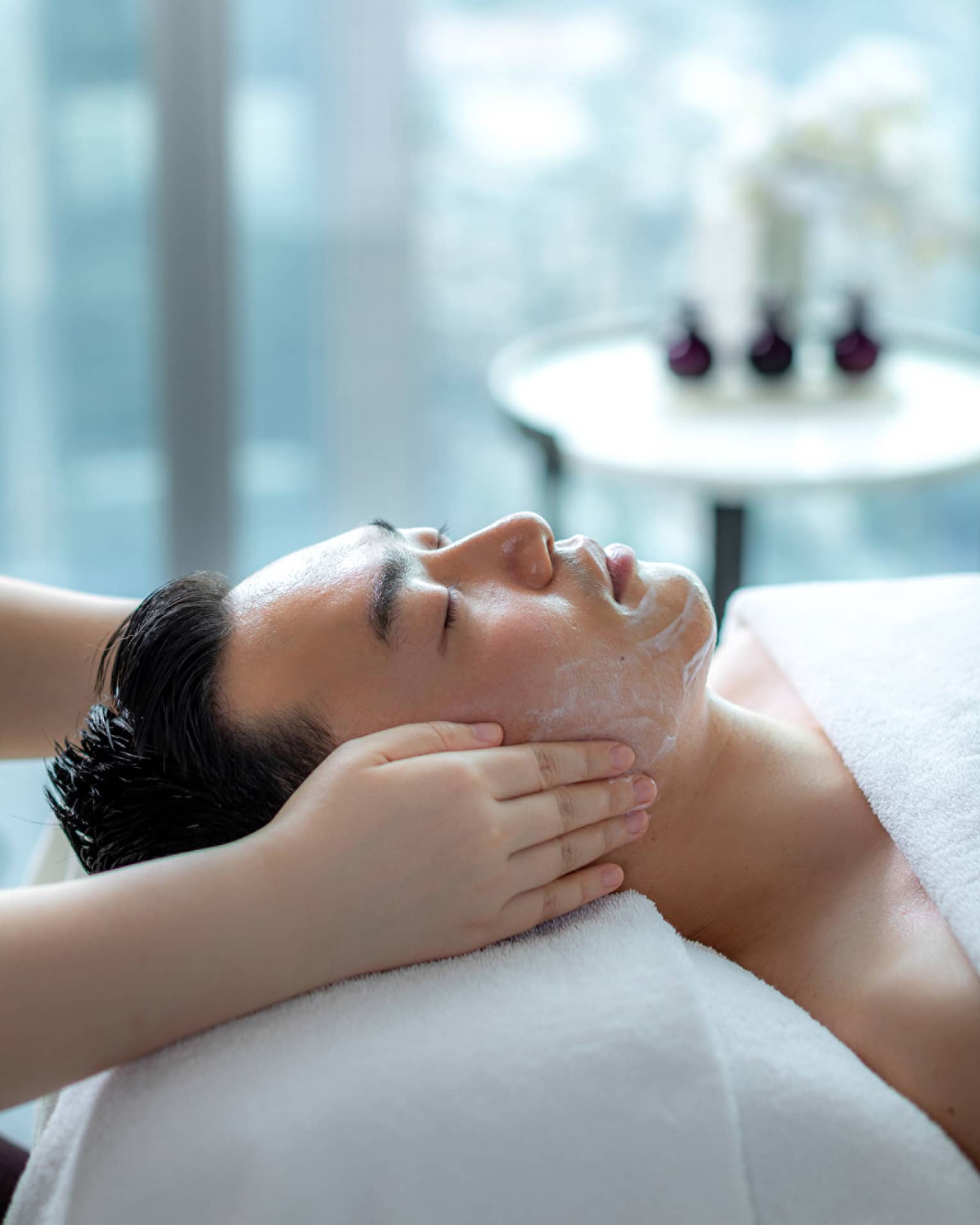 Hands rest on man's cheeks as he lays on massage table by glass window with daytime city view 