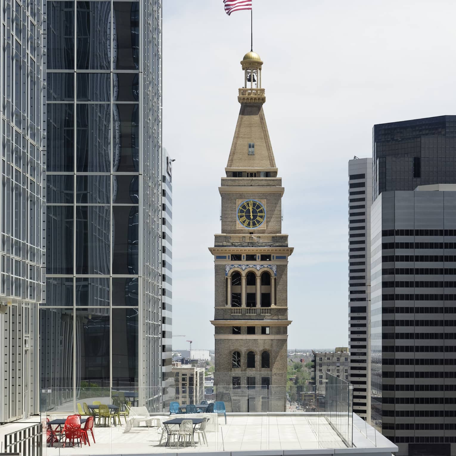 Denver's distinctive, historic clock tower, American flag on top, rises above a top floor patio amid modern skyscrapers.