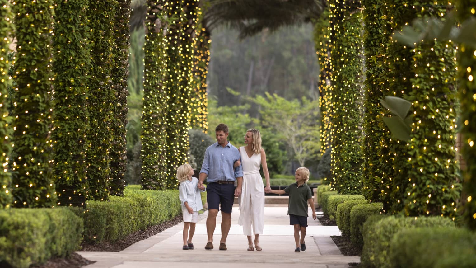 A mother, father and two sons walking along a path of palm trees wrapped in Christmas lights.