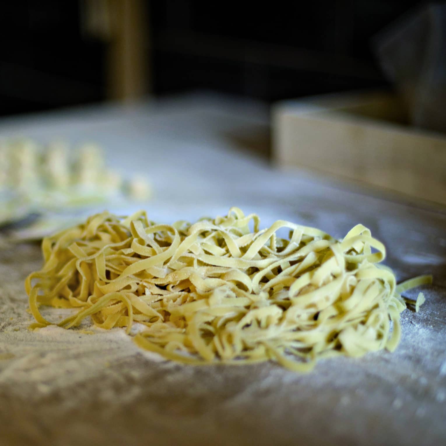 Pile of fresh tagliatelle rests on an aged wood counter covered with flour, beside scraps of pasta dough. 