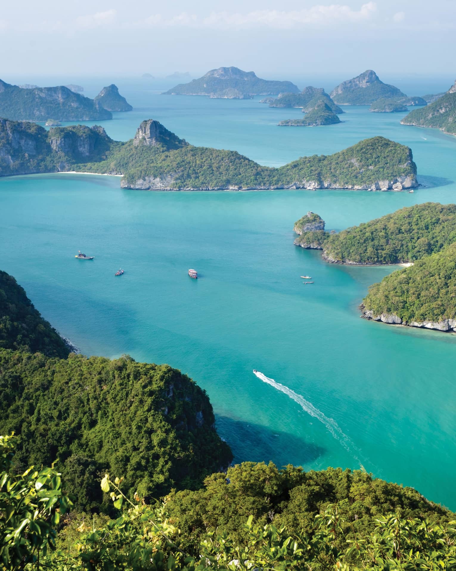 Aerial view of a dozen lush, forested, hilly islands of different sizes surrounded by calm turquoise water dotted with boats.