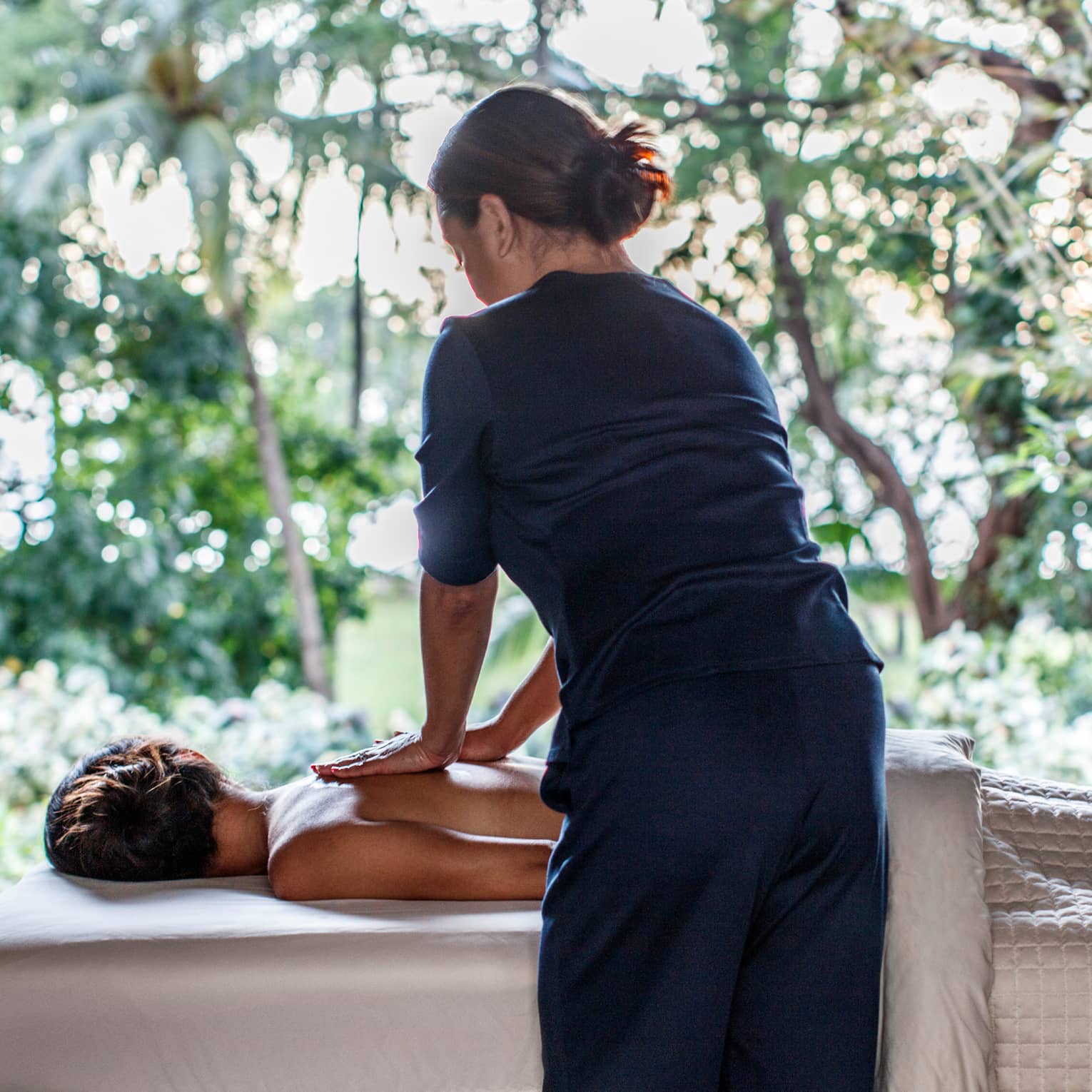 Spa staff massages woman's bare shoulders as she lies under sheet on massage table