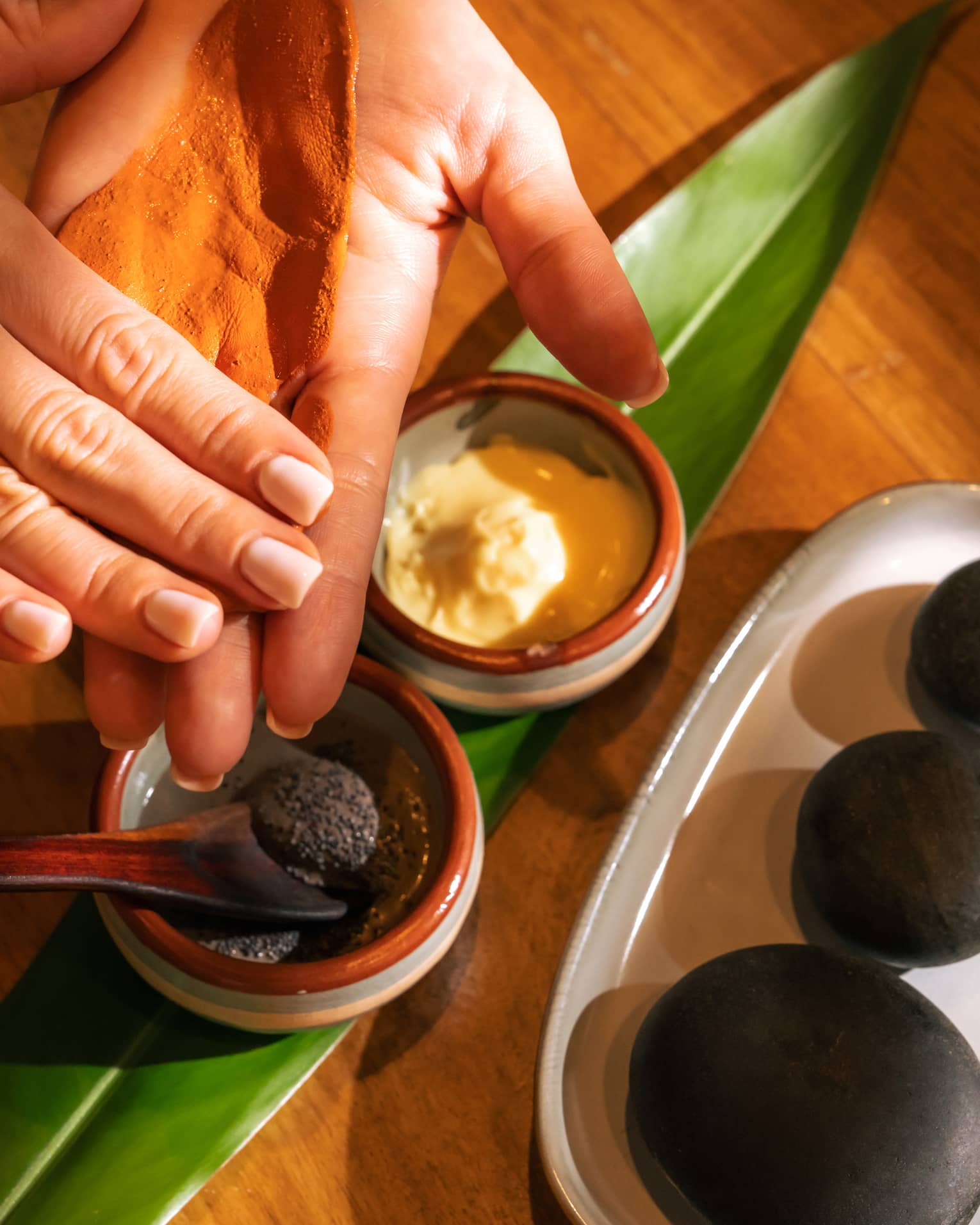 Pair of hands tests spa ingredients from two small stoneware bowls set on a fresh green leaf with three black stones placed on a dish nearby