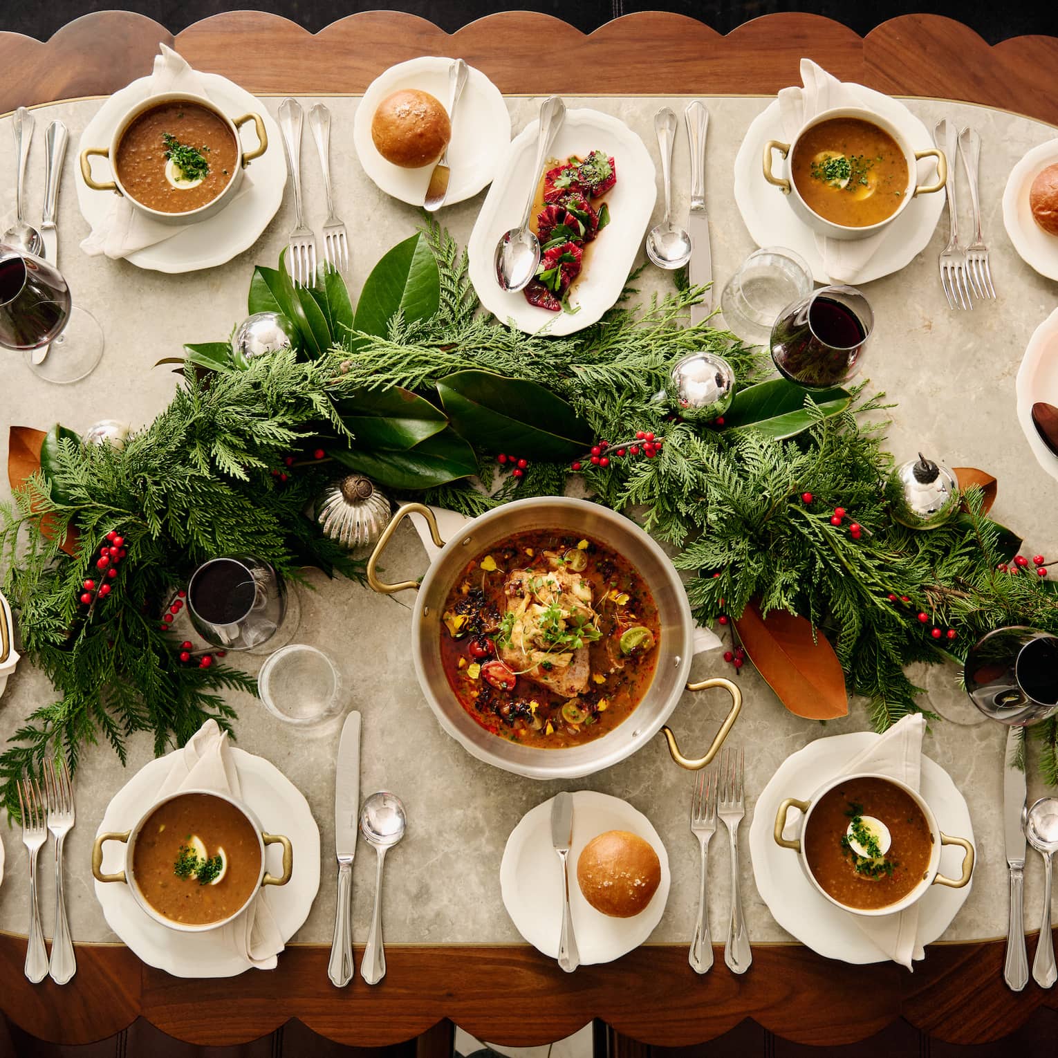 View from above of hands placing a dish on a table laden with soups, stews, bread and wine and adorned with festive greenery.