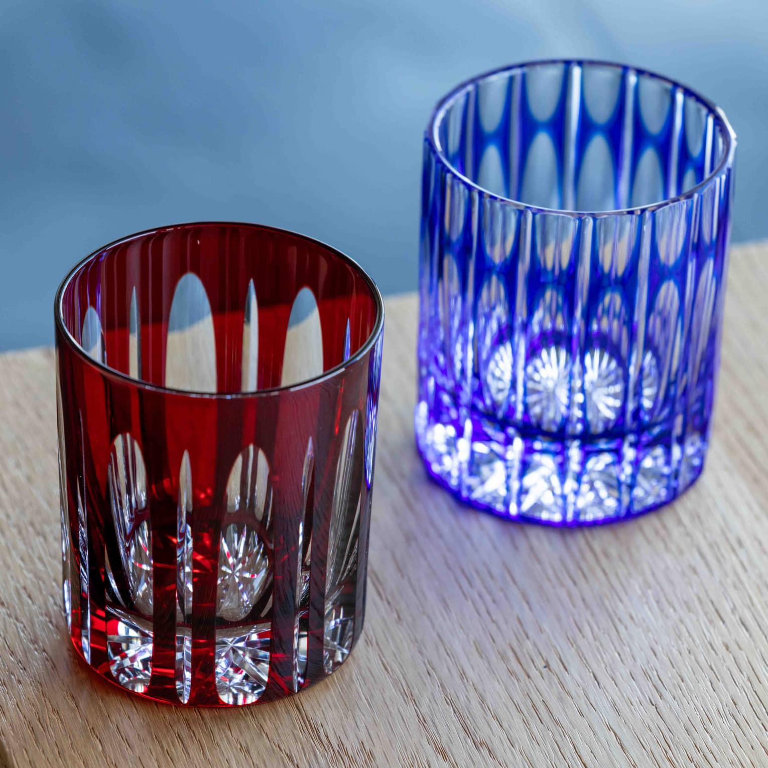 Close-up of two cut-glass tumblers with intricate geometric patterns overlaid by coloured glass, one dark red, one blue.