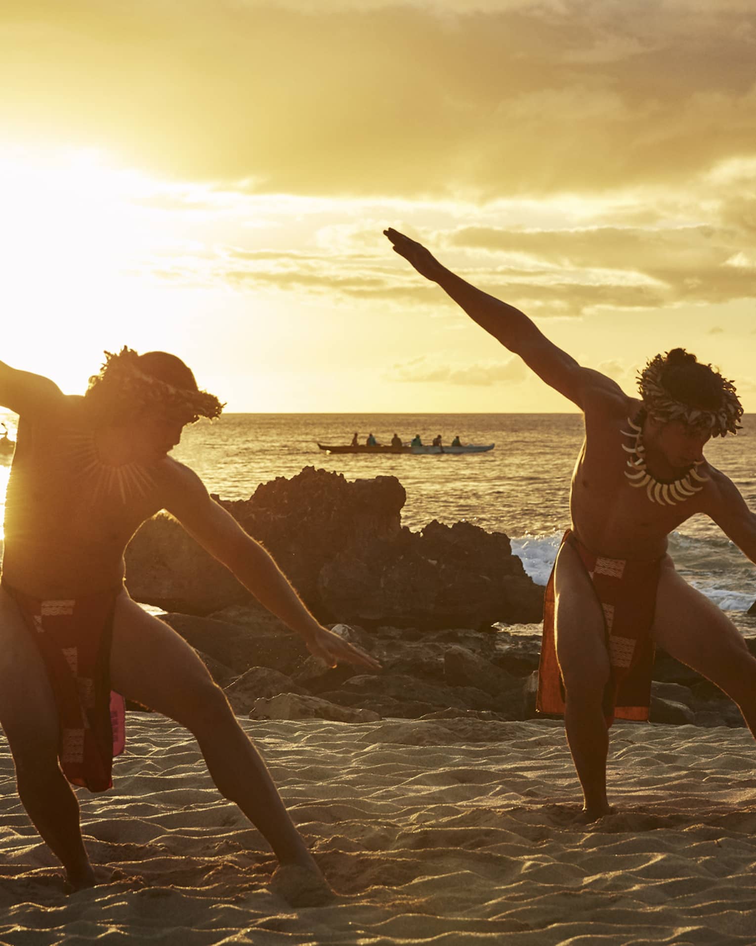 Two Hawaiian dancers with arms outstretched pose on sandy beach at sunset