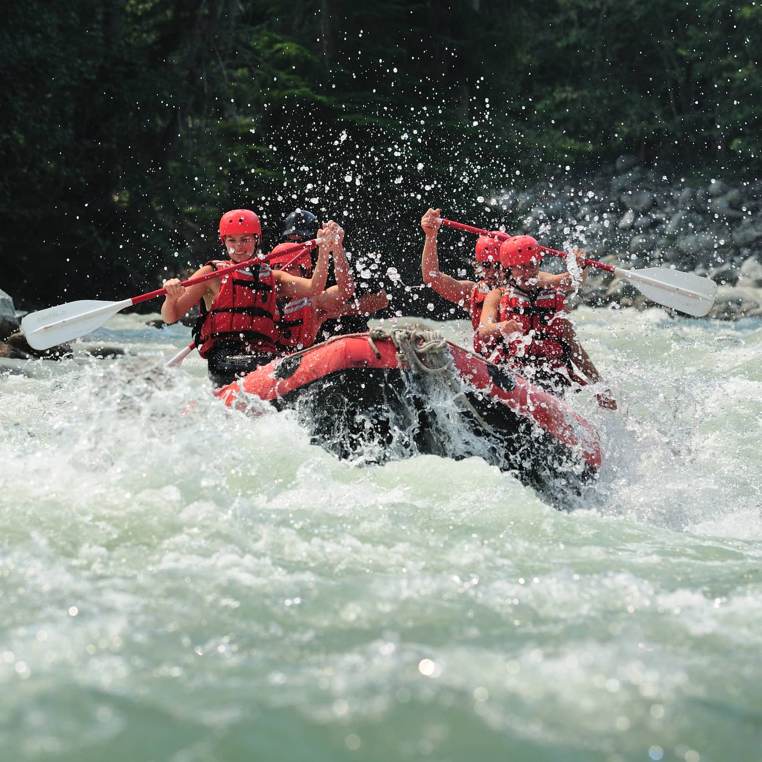 Smiling paddlers in red helmets and lifejackets steer an inflated raft through raging white water, passing evergreen trees.