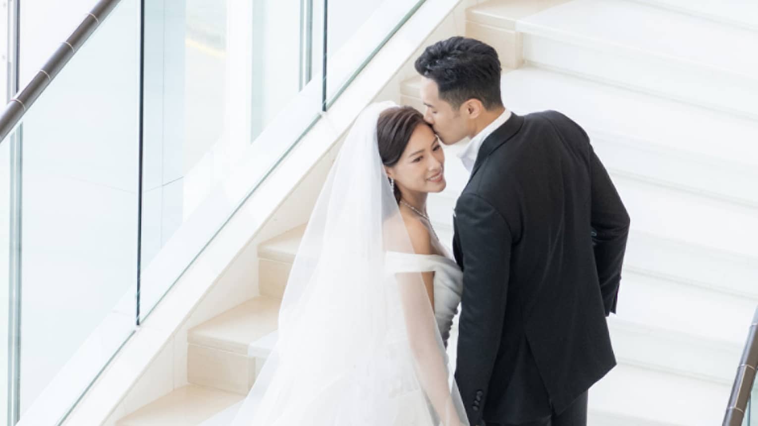 A bride and groom walking up stairs near large windows.