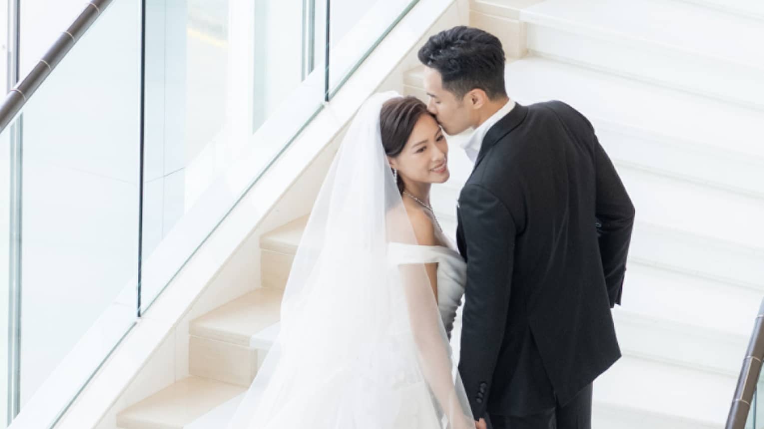 A bride and groom walking up stairs near large windows.