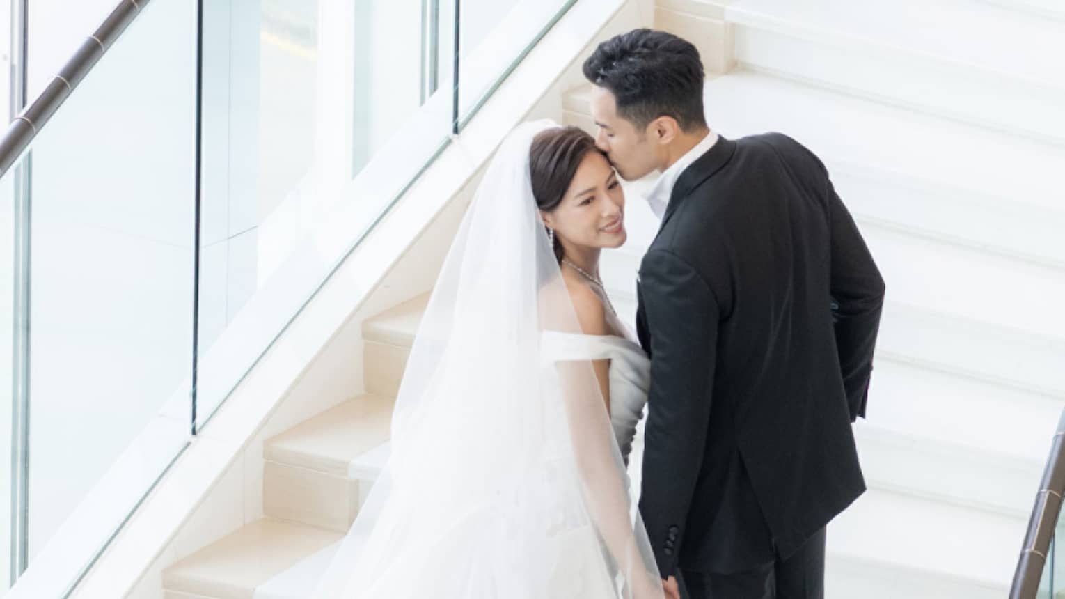 A bride and groom walking up stairs near large windows.