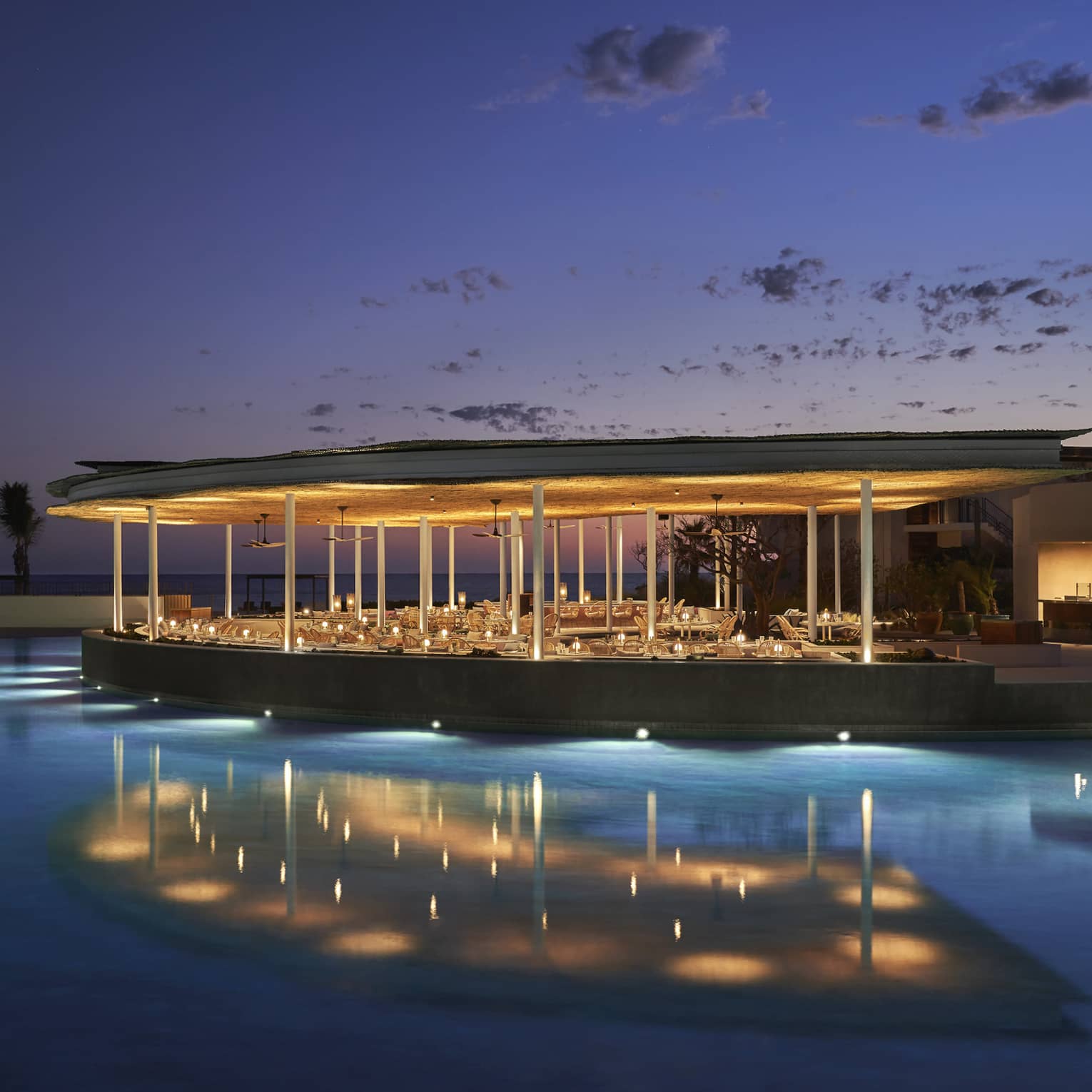 The exterior of an outdoor eating area at night on the water of a shore, it is well lit with lights.