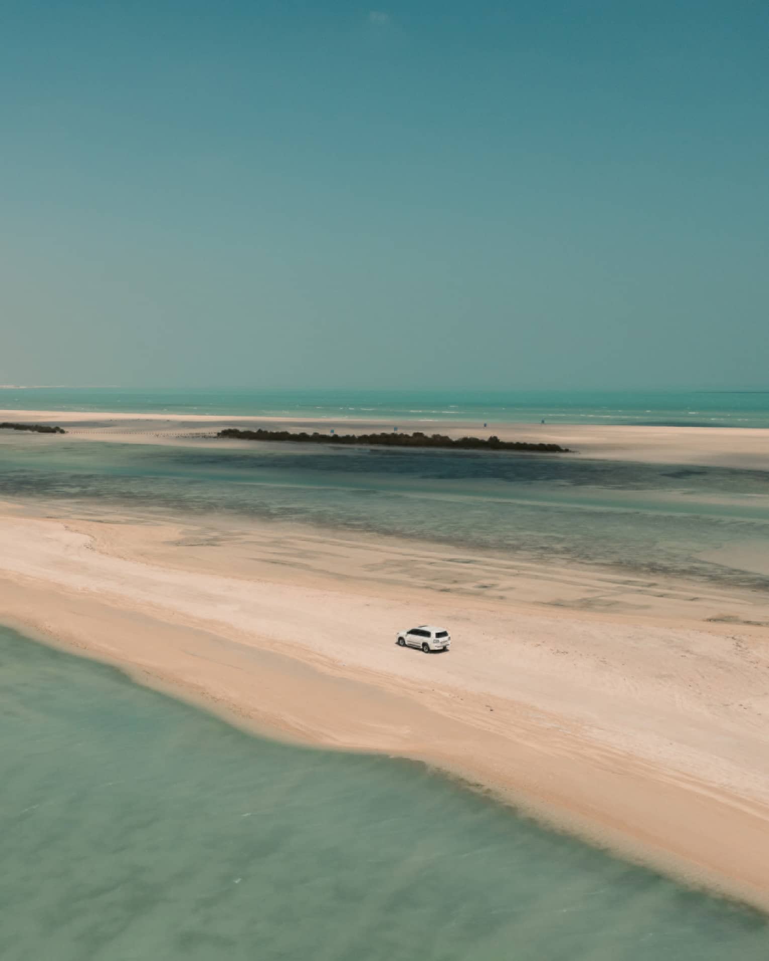 Aerial view of a 4x4 car traversing a sandbar surrounded by shallow turquoise water on either side and a cloudless sky above.